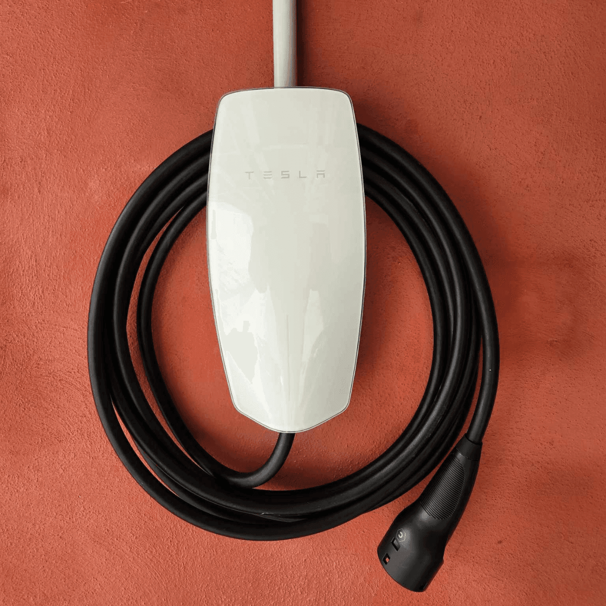 Tesla Gen 3 Wall Connector EV charger with a glossy white front panel and black coiled charging cable, mounted on a textured orange wall.