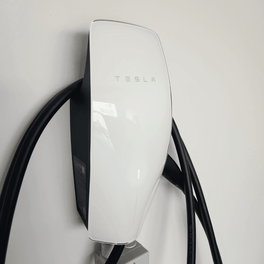 Tesla Gen 3 Wall Connector EV charger mounted on a white interior wall with a sleek, glossy white front panel displaying the Tesla logo, thick black charging cable, and green indicator light